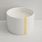 White candle in a bowl with a gold accent and logo on a light gray background