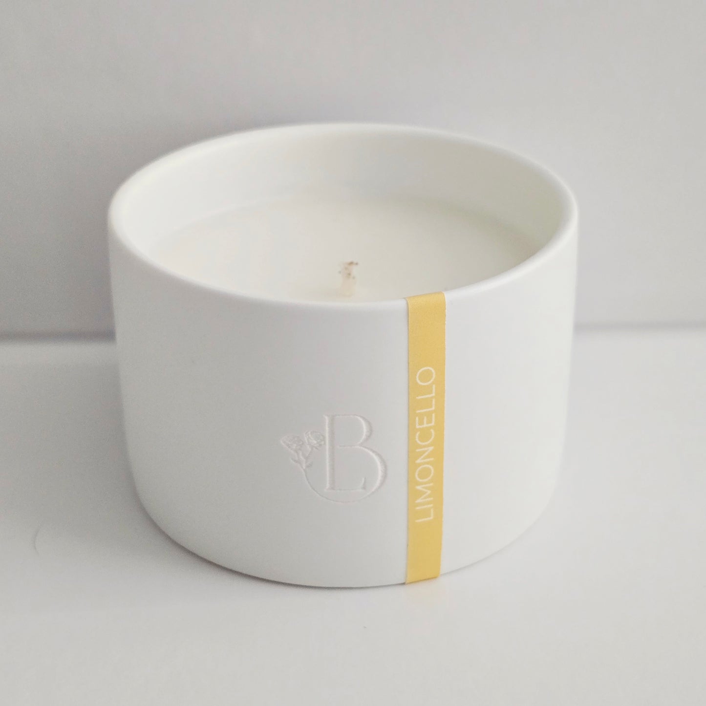 White candle in a bowl with a gold accent and logo on a light gray background