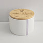 White candle with wooden lid labeled 'Love Blooms' on a light gray background