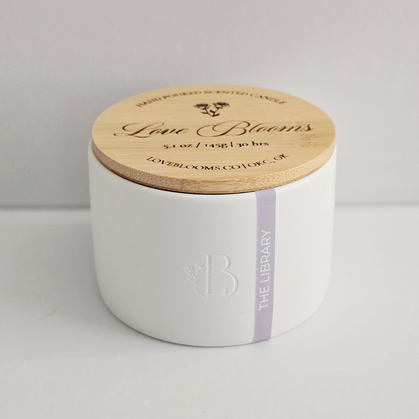 White candle with wooden lid labeled 'Love Blooms' on a light gray background