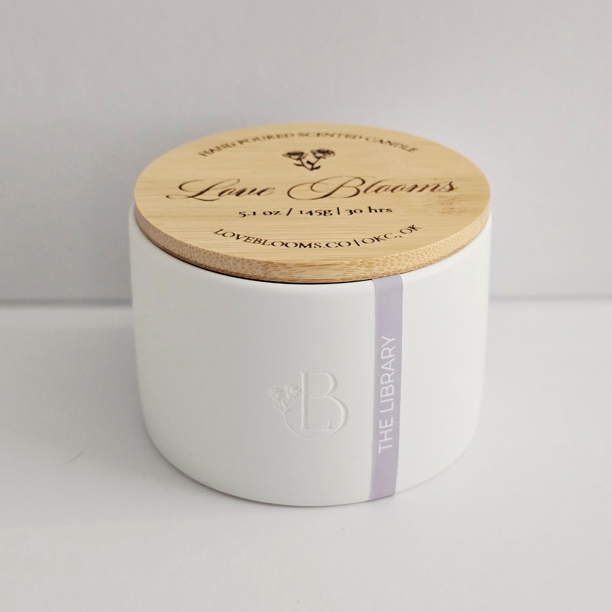 White candle with wooden lid labeled 'Love Blooms' on a light gray background
