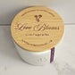 White candle with wooden lid labeled 'Love Blooms' on a light background