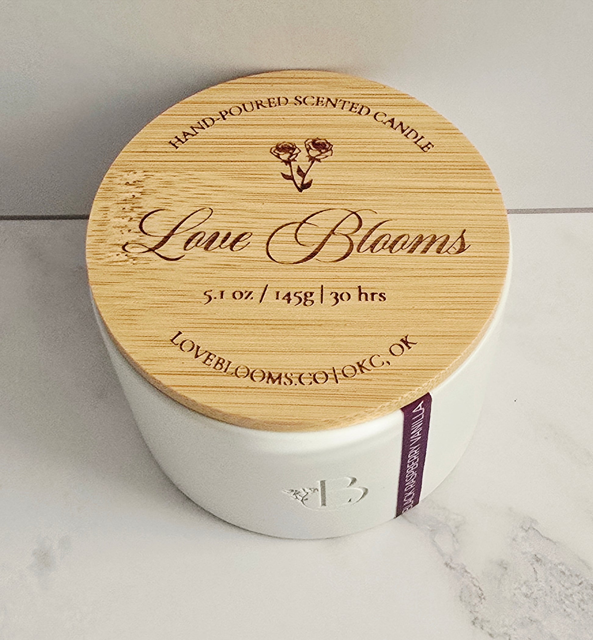 White candle with wooden lid labeled 'Love Blooms' on a light background