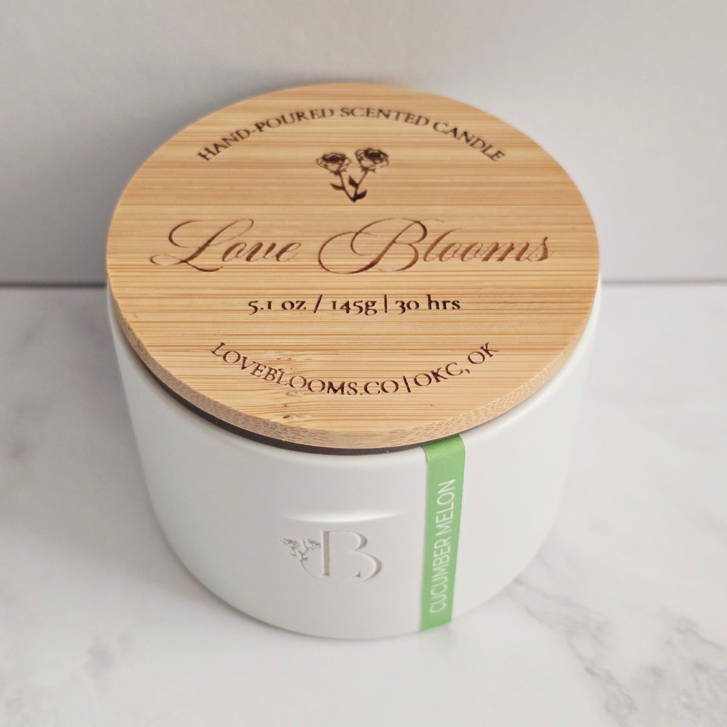 White candle with a wooden lid labeled 'Love Blooms' on a light background